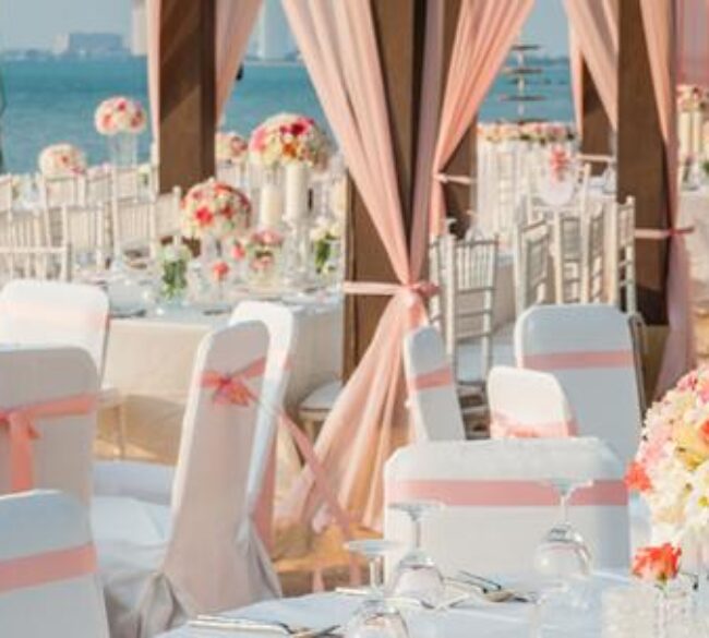 Outdoor wedding reception setup with round tables covered in white tablecloths and pink sashes, floral centerpieces, white chairs, and a waterfront view in the background.
