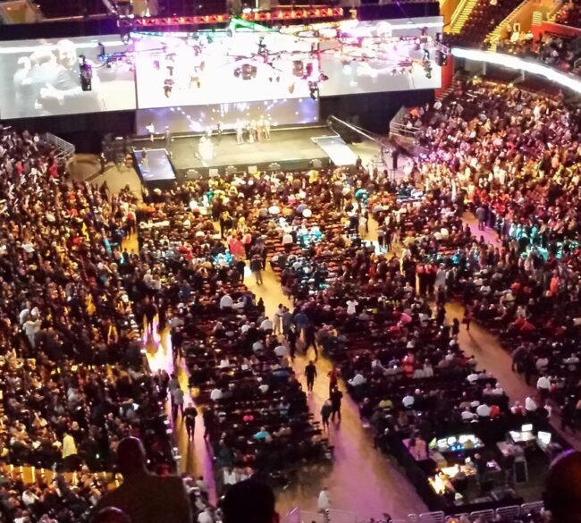 A wide-angle view of a full indoor arena with a stage and large screens in the center demonstrating how Prismm can help create immersive experiences for any venue — before the event even begins.