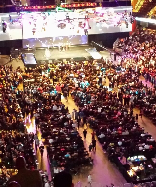 A wide-angle view of a full indoor arena with a stage and large screens in the center demonstrating how Prismm can help create immersive experiences for any venue — before the event even begins.