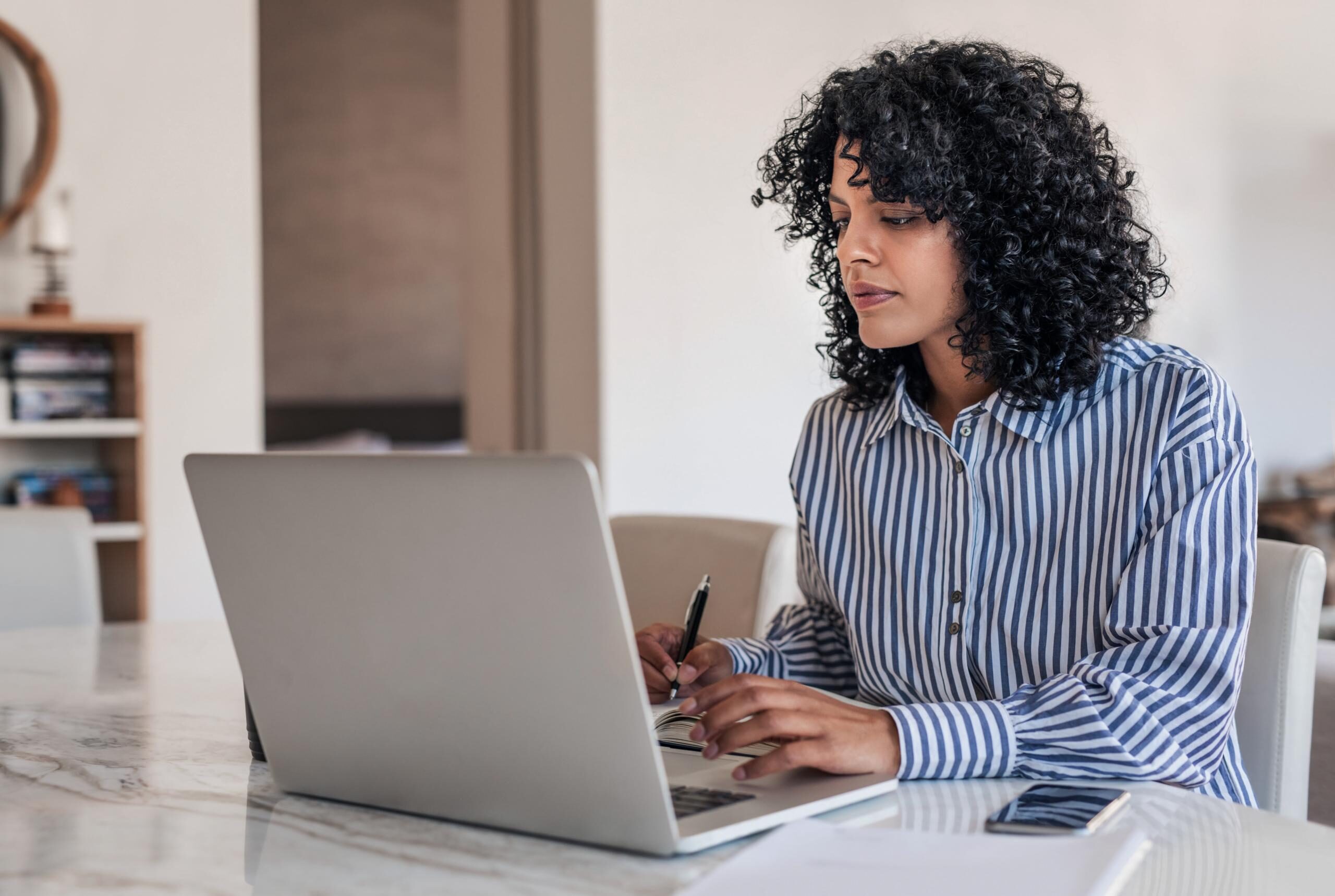 Young female entrepreneur using a laptop and writing down notes while working from home at her dining table